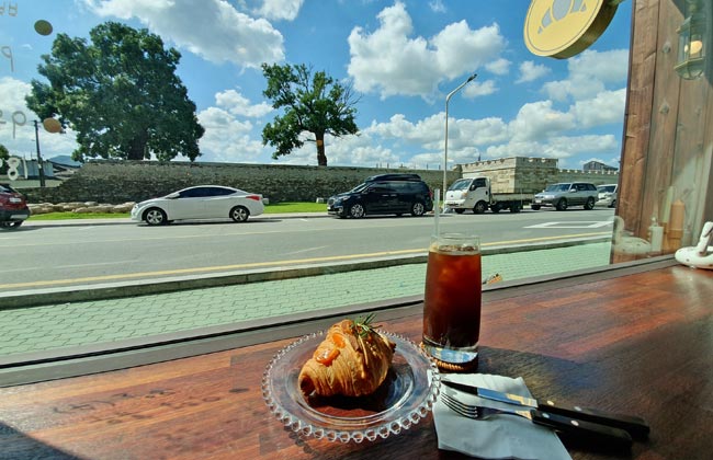 테이블 위로 커피와 크루아상이 놓여있고 창밖으로 경주읍성이 보이는 사진