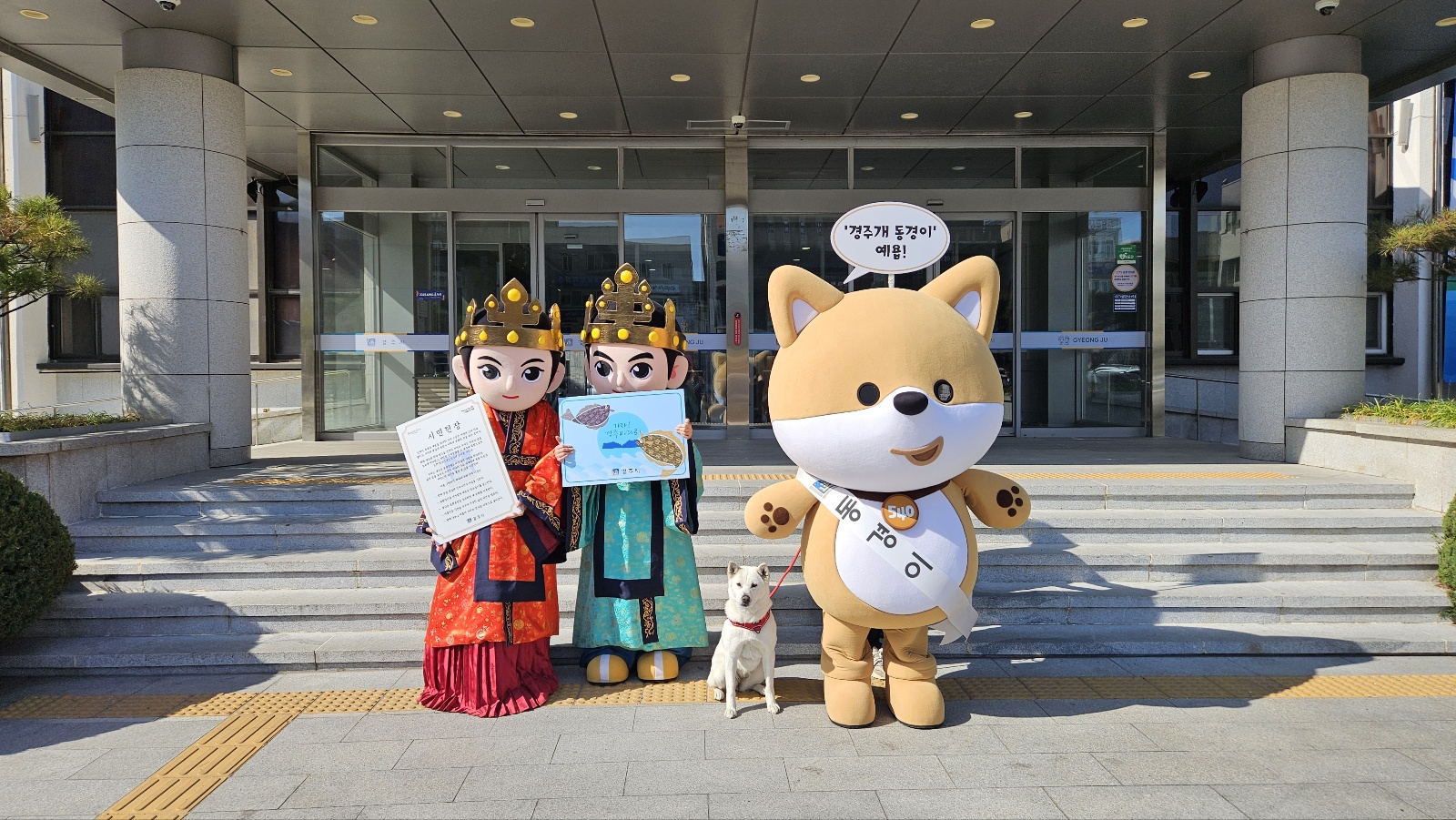경주시청 본관 앞에서 경주시 공식 상징물로 지정된 캐릭터 ‘관이’(왼쪽), ‘금이’(가운데), ‘동경이’(오른쪽)와 천연기념물 제540호 ‘경주개 동경이’가 시민헌장과 참가자미 캐릭터 홍보물을 들고 포즈를 취하고 있다.